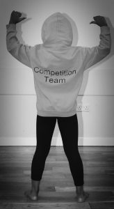 a person in a hoodie with competition teams printed on the back faces away from the camera, pointing their raised thumbs towards themselves